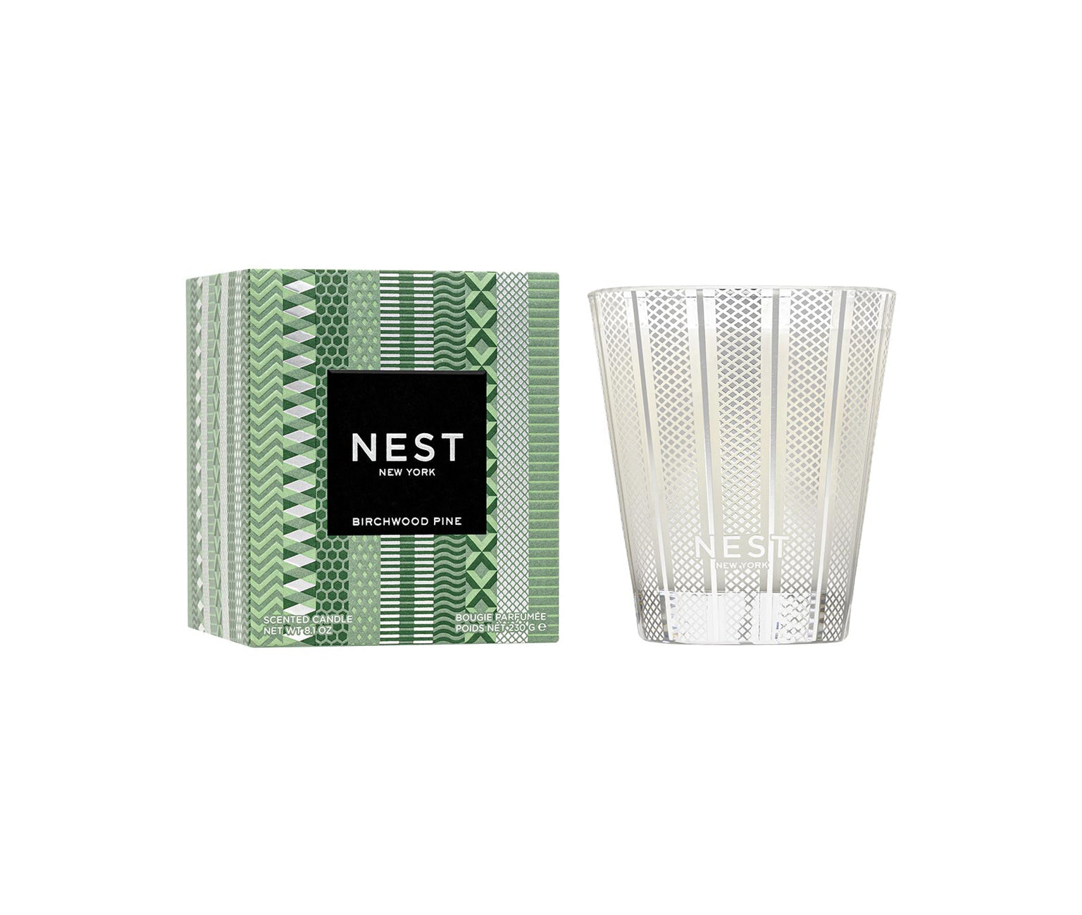 The Nest Birchwood Pine Collection candle, in clear patterned glass beside its green box with a black-and-white label, fills your space with a winter forest scent for a festive atmosphere.