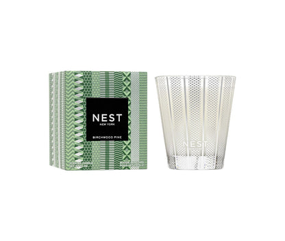 The Nest Birchwood Pine Collection candle, in clear patterned glass beside its green box with a black-and-white label, fills your space with a winter forest scent for a festive atmosphere.