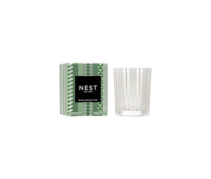 The Nest Birchwood Pine Collection holiday candle, shown in a clear, patterned glass jar beside its green and white box, evokes the crisp fragrance of a winter forest.