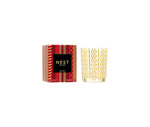 A Nest Holiday Collection candle in a gold-patterned glass holder with a festive scent is displayed next to its decorative red and gold box.