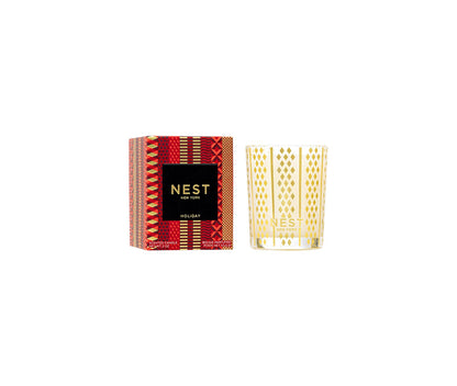A Nest Holiday Collection candle in a gold-patterned glass holder with a festive scent is displayed next to its decorative red and gold box.