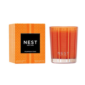 A Nest Pumpkin Chai Candle Collection candle in orange patterned glass sits beside its matching box, both featuring elegant black labels, creating the perfect autumn ambiance.
