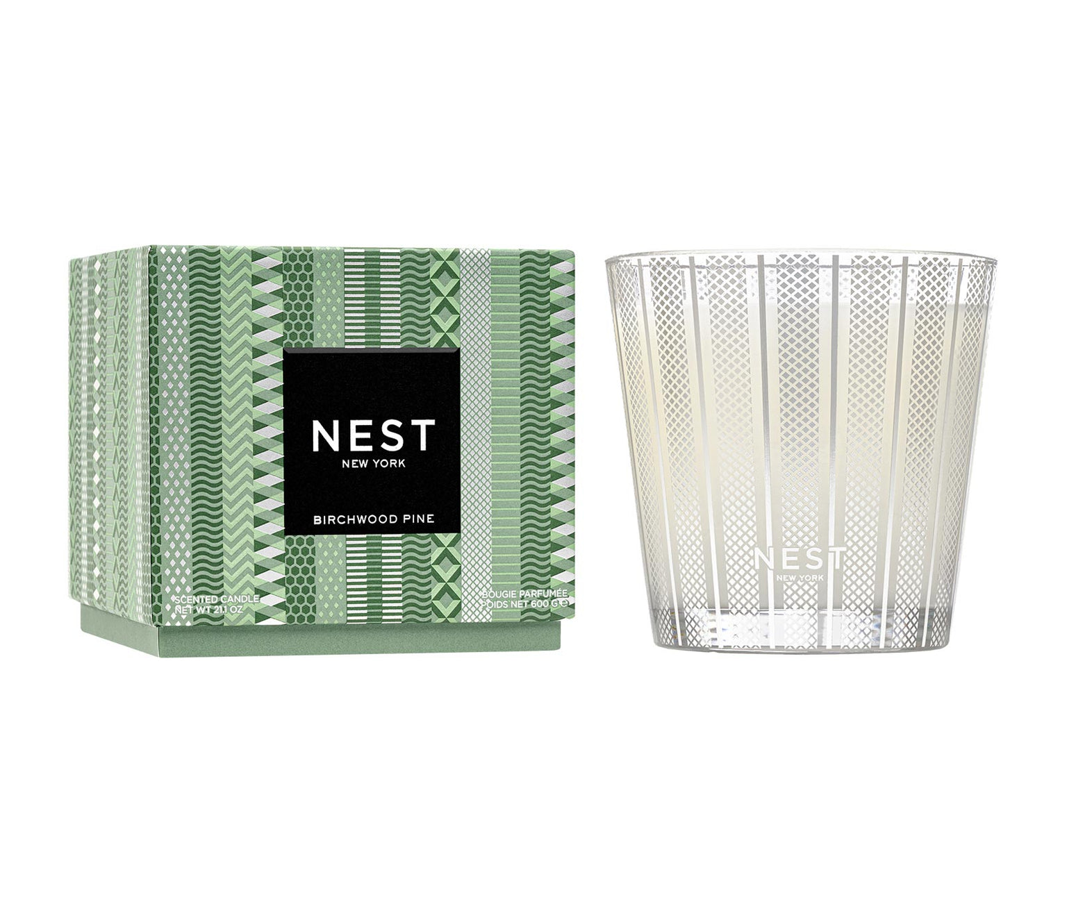 A Nest Birchwood Pine Collection holiday candle in a patterned glass sits beside its green and white box, evoking the fresh, wintry scent of a forest.