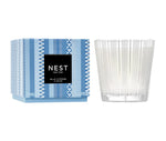 A Nest Blue Cypress & Snow Collection candle in a patterned blue box (left) and clear textured glass holder (right), offering a winter scent with smoked vanilla bean notes.