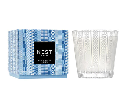 A Nest Blue Cypress & Snow Collection candle in a patterned blue box (left) and clear textured glass holder (right), offering a winter scent with smoked vanilla bean notes.