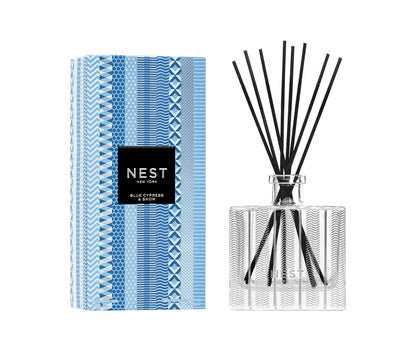 The Nest Blue Cypress & Snow Collection reed diffuser features a patterned glass bottle with black reeds and comes in a blue and white geometric box—ideal for adding a cozy winter scent to your space.