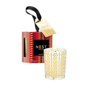 A small NEST Holiday Collection candle in a patterned glass jar sits next to its decorative red and gold box with an attached gold gift tag.