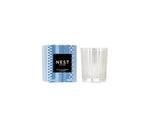 A Nest Blue Cypress & Snow Collection candle with smoked vanilla bean winter scent, in a clear glass holder next to its blue and white patterned box featuring a black label.