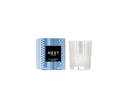 A Nest Blue Cypress & Snow Collection candle with smoked vanilla bean winter scent, in a clear glass holder next to its blue and white patterned box featuring a black label.