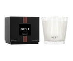 A Nest Something Tropical Candle from the Candle and Diffuser Collection is displayed in a glass holder beside its black box, suggesting a blend of passion fruit and cashmere woods.