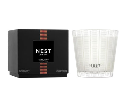 A Nest Something Tropical Candle from the Candle and Diffuser Collection is displayed in a glass holder beside its black box, suggesting a blend of passion fruit and cashmere woods.