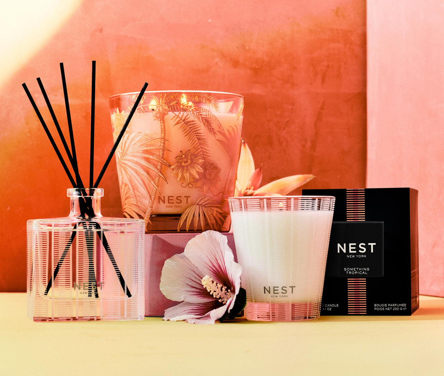 The Nest Something Tropical Candle and Diffuser Collection features candles with passion fruit notes, a reed diffuser, a product box, and a pink hibiscus flower, all set against a vibrant pink and yellow background.