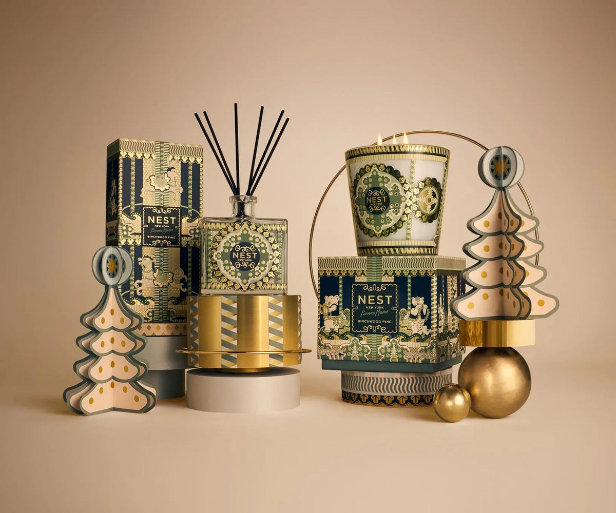 Pierre Marie Birchwood Collection home fragrance products, including a reed diffuser, 3-wick candle, and boxed items, arranged with gold-toned geometric and tree-shaped ornaments.