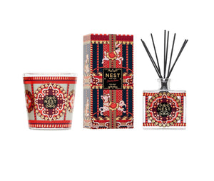A Nest Pierre Marie Holiday Collection pomegranate pine 3-wick candle in a patterned glass jar, a decorative box, and a square reed diffuser with ornate red, gold, and blue holiday-themed designs.