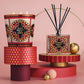 A Nest Pierre Marie Holiday Collection 3-wick pomegranate pine candle and a reed diffuser with intricate red, gold, and black patterned labels, displayed on tiered geometric platforms.