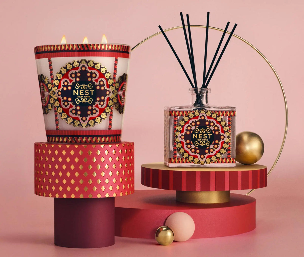 A Nest Pierre Marie Holiday Collection 3-wick pomegranate pine candle and a reed diffuser with intricate red, gold, and black patterned labels, displayed on tiered geometric platforms.