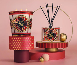 A Nest Pierre Marie Holiday Collection 3-wick pomegranate pine candle and a reed diffuser with intricate red, gold, and black patterned labels, displayed on tiered geometric platforms.