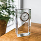 The Simon Pearce Woodbury Tall Clock, with its rectangular glass body and silver-rimmed round face, rests on a wooden surface; a potted plant is softly blurred in the background.