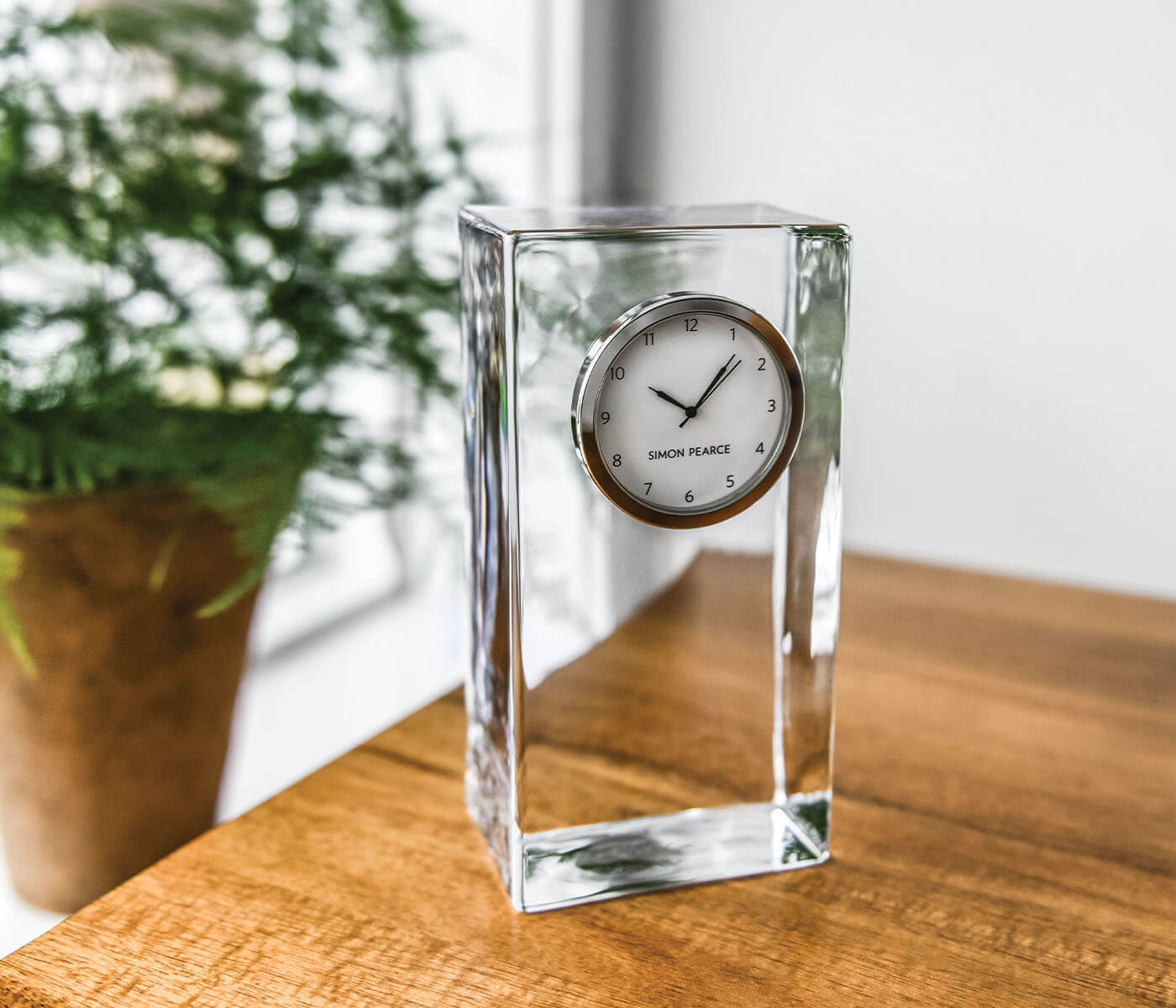 The Simon Pearce Woodbury Tall Clock, with its rectangular glass body and silver-rimmed round face, rests on a wooden surface; a potted plant is softly blurred in the background.