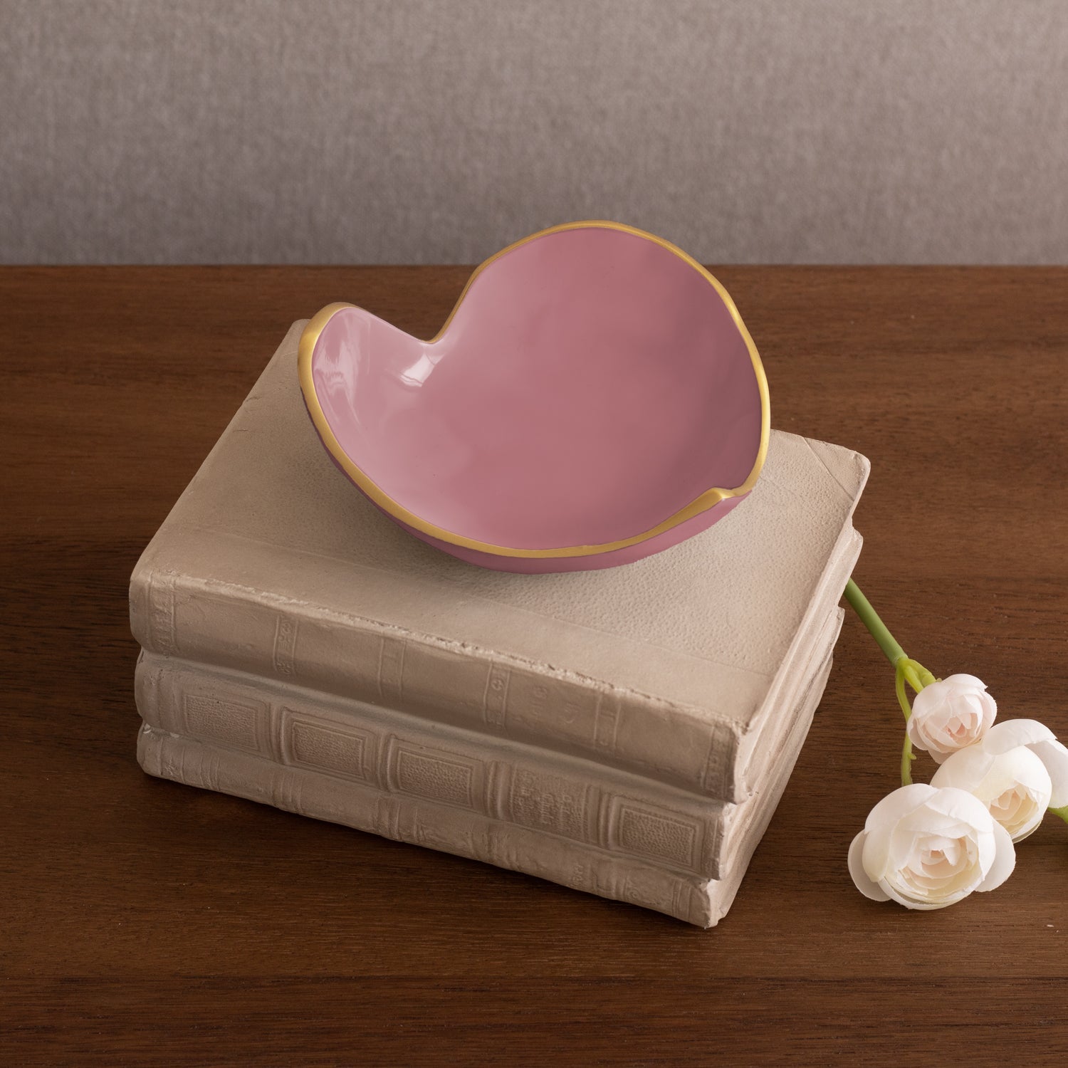 A pink, gold-rimmed heart dish from the Beatriz Ball Encanto Soho Mini Heart Dish Collection sits on three beige books beside two white artificial flowers on a wooden surface.
