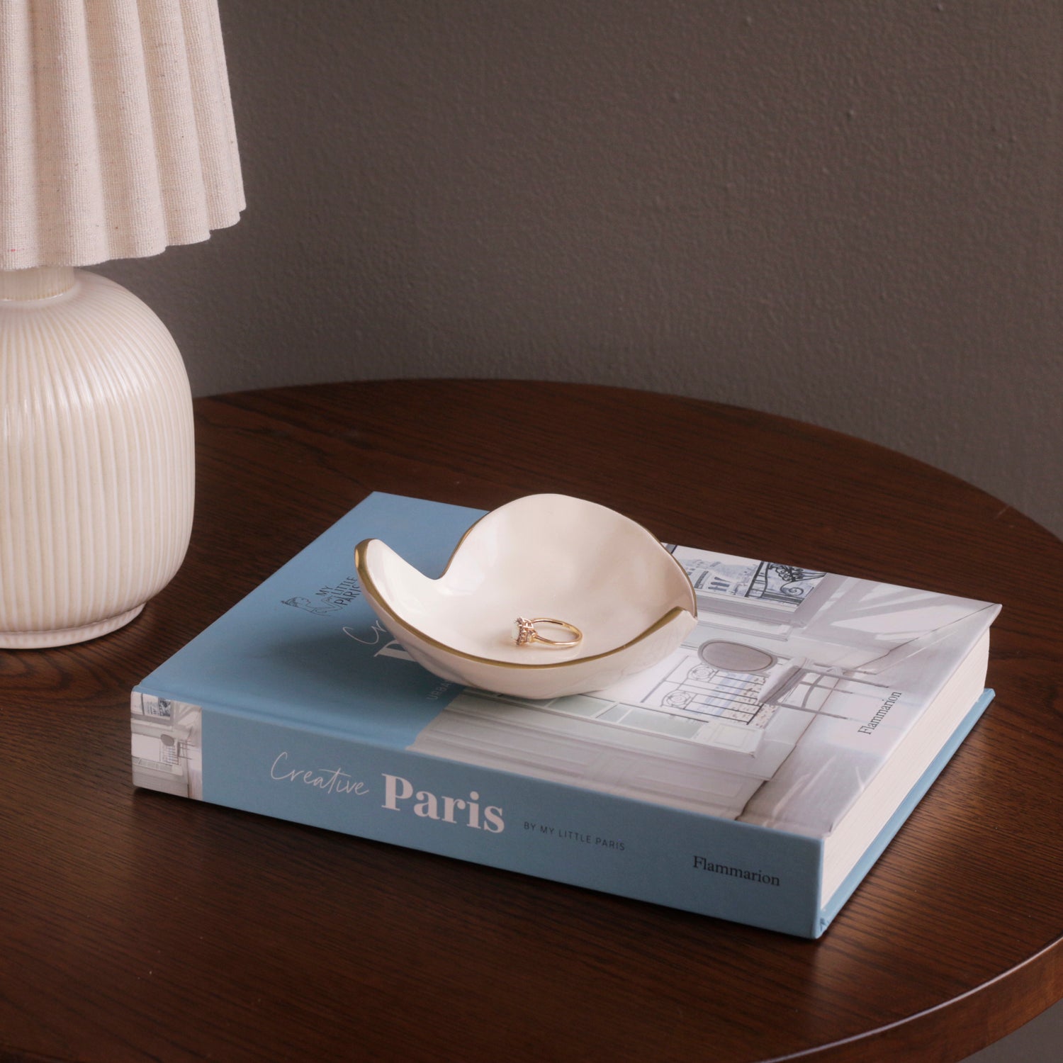 A Beatriz Ball Encanto Soho Mini Heart Dish from the multicolored collection holds a ring atop a closed "Creative Paris" book, next to a white table lamp on wood—a charming setting for a handmade gift.