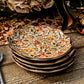Four Willa Dinnerware Collection plates by Juliska, featuring romantic florals and an autumn leaf pattern, are stacked on a wooden table with flowers and a patterned tablecloth in the background.
