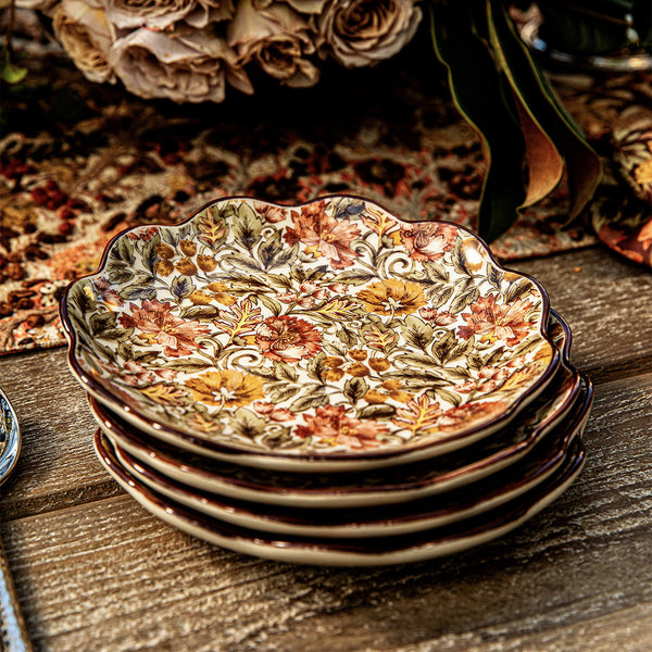 Four Willa Dinnerware Collection plates by Juliska, featuring romantic florals and an autumn leaf pattern, are stacked on a wooden table with flowers and a patterned tablecloth in the background.
