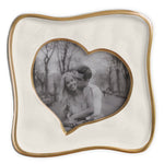 A handcrafted 5x5 Encanto Curved Heart Frame by Beatriz Ball displays a black-and-white photo of a smiling couple and features a wavy gold border, abstract heart shape, and charming trees and path in the background.