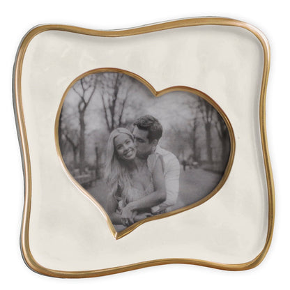 A handcrafted 5x5 Encanto Curved Heart Frame by Beatriz Ball displays a black-and-white photo of a smiling couple and features a wavy gold border, abstract heart shape, and charming trees and path in the background.
