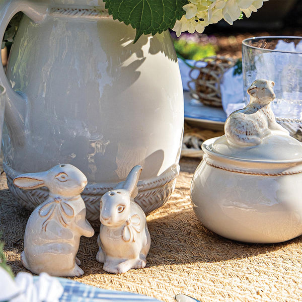 Juliska’s Whitewash Bunny Serveware Collection, featuring ceramic bunny figurines and a lidded jar with an animal figure, is arranged on a woven mat alongside a large pitcher, glassware, and charming ceramic tableware.