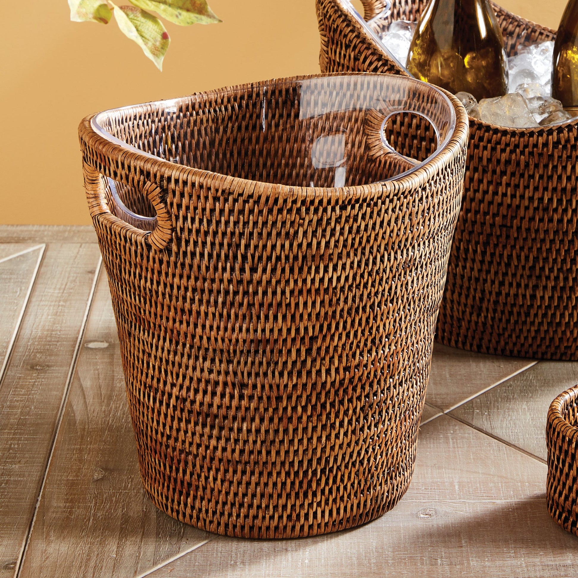 The Napa Home & Garden Burma Rattan Beverage Tub, featuring two side handles and a watertight design, rests on a wooden surface. Another similar basket and objects are partially visible in the background.