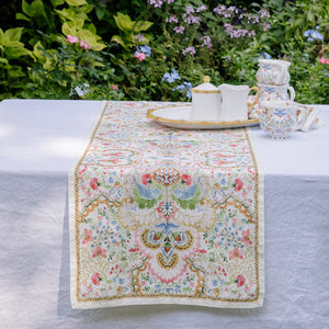 A white table is adorned with the Juliska Sofia Table Runner and an ornate tea set, while green foliage and blooming flowers in the background create a charming spring table decor.