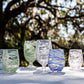 Six glasses from the Juliska Puro Marbled Drinkware Collection, hand-blown with swirling green, clear, and blue hues, are displayed on a white surface outdoors with trees and sunlight in the background.