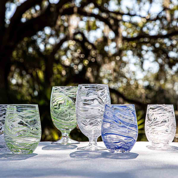 Six glasses from the Juliska Puro Marbled Drinkware Collection, hand-blown with swirling green, clear, and blue hues, are displayed on a white surface outdoors with trees and sunlight in the background.