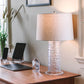 A handblown glass table lamp with a beige shade sits on a wooden desk alongside a closed laptop, two tablets, potted plants by a window, and the Simon Pearce Barre Clock.