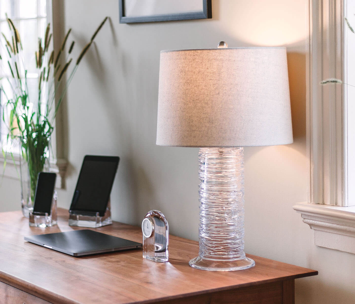 A handblown glass table lamp with a beige shade sits on a wooden desk alongside a closed laptop, two tablets, potted plants by a window, and the Simon Pearce Barre Clock.
