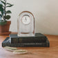 The Simon Pearce Barre Clock, a handblown glass timepiece, sits atop two stacked books on a wooden surface with a potted plant in the background—adding a modern touch to any space.
