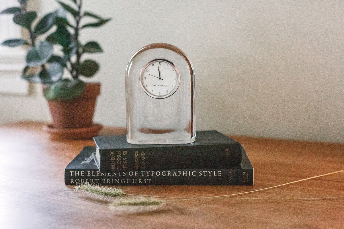 The Simon Pearce Barre Clock, a handblown glass timepiece, sits atop two stacked books on a wooden surface with a potted plant in the background—adding a modern touch to any space.