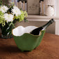 A Beatriz Ball Encanto Leaf Green & White Serveware Collection ceramic ice bucket holds champagne on a wooden table, with matching enameled bowls, a vase of white flowers, and books in the background.