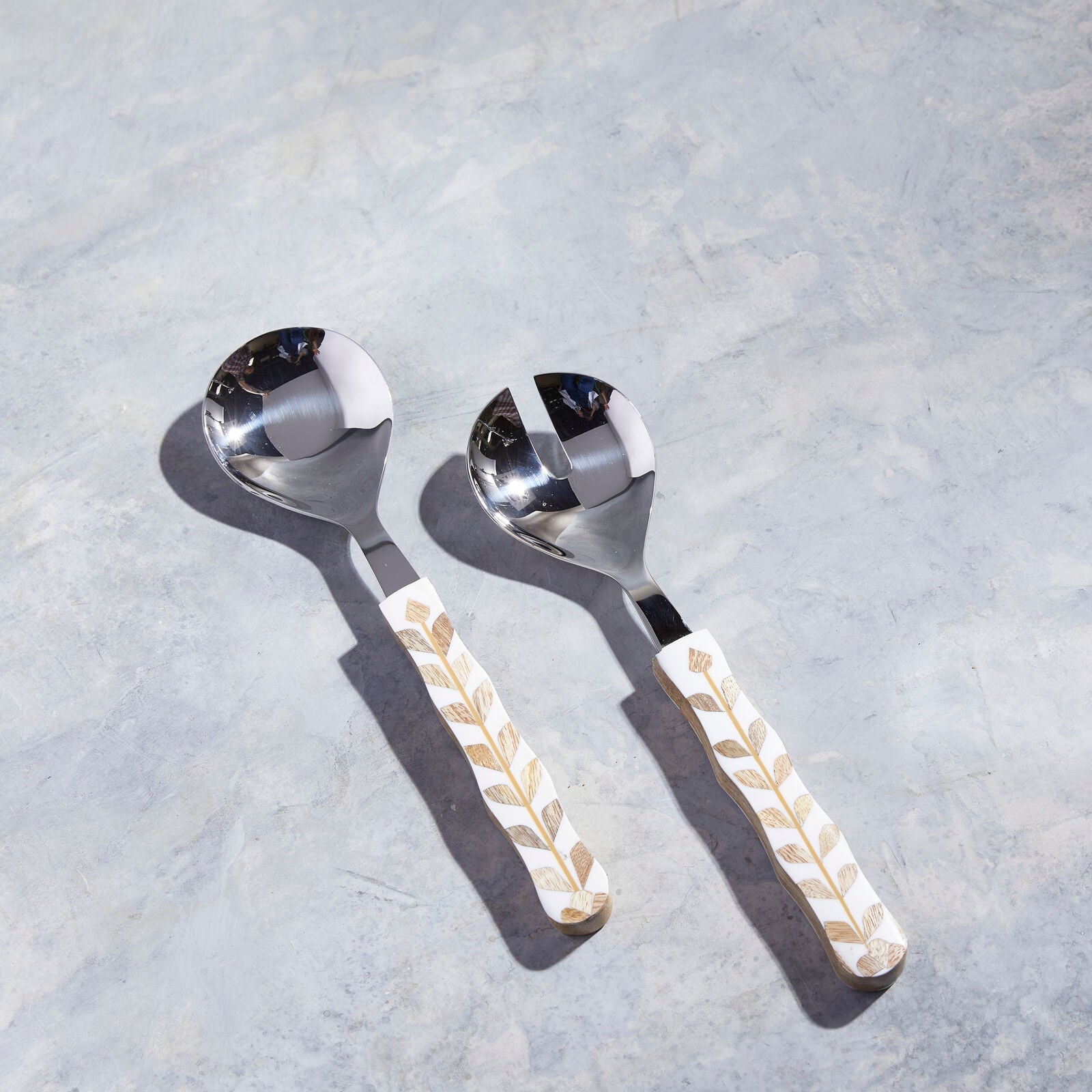 Two metal serving spoons from the Mya Inlay Serving and Decor Collection by Be Home, featuring white and gold leaf-patterned handles with mother of pearl accents, displayed on a light gray textured surface.