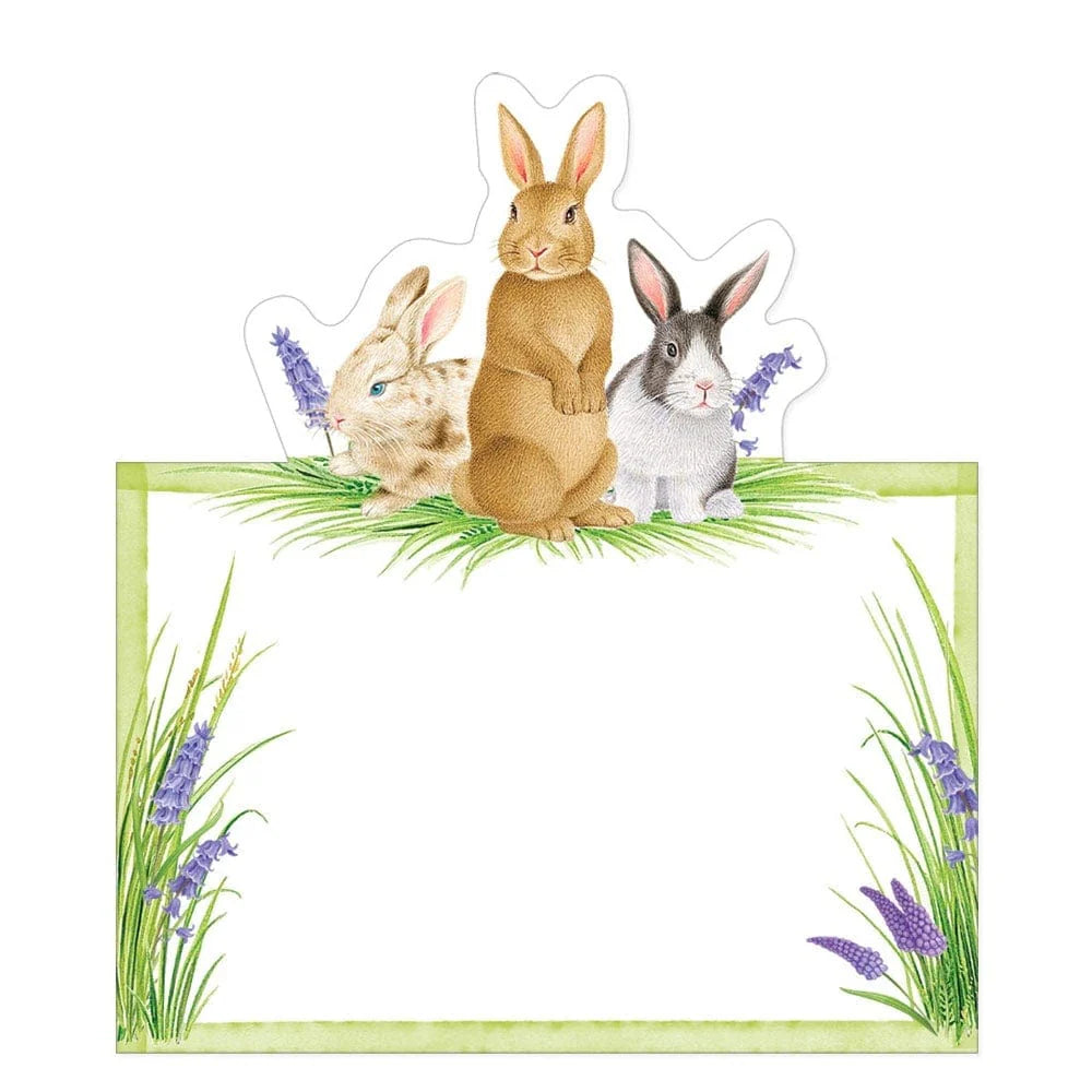 Three illustrated rabbits from Caspari’s Bunnies and Daffodils Place Setting Collection perch on a blank sign edged with green grass and purple flowers—perfect Easter or spring party decor.