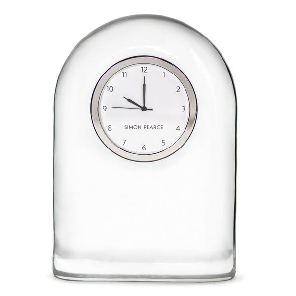 The Simon Pearce Barre Clock features a clear handblown glass design with a white face and black hands set at 12:15. Marked hours enhance readability, complementing its contemporary style. The label reads "Simon Pearce.