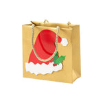 The Be Merry Collection gold gift bag by Caspari features rope handles and a festive front with a red Santa hat and holly berry—perfect for holiday gifting with its standout design.