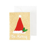The Caspari "Be Merry Collection" greeting card features a red Santa hat, holly leaves, and "Be Merry" on a beige background, paired with a white envelope.
