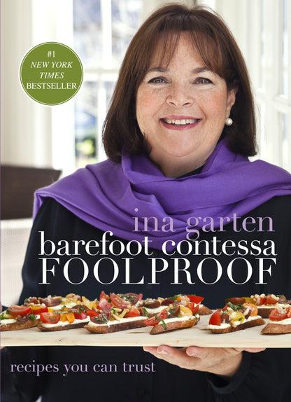 A woman with shoulder-length brown hair and a purple scarf holds a tray of appetizers, including Hot Smoked Salmon; the book “Barefoot Contessa Foolproof” by Penguin Random House is visible on the cover, authored by Ina Garten.