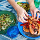 Person's hands arranging seafood on a blue plate, accompanied by a green dish with vegetables, a blue glass, and a bottle on a blue-patterned tablecloth—perfect for impromptu dinner ideas that are visually stunning. Featuring Nothing Fancy: The Art of Having People Over by Common Ground.