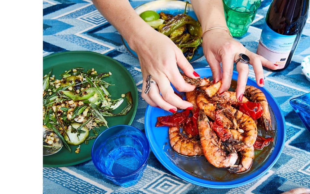 Person's hands arranging seafood on a blue plate, accompanied by a green dish with vegetables, a blue glass, and a bottle on a blue-patterned tablecloth—perfect for impromptu dinner ideas that are visually stunning. Featuring Nothing Fancy: The Art of Having People Over by Common Ground.