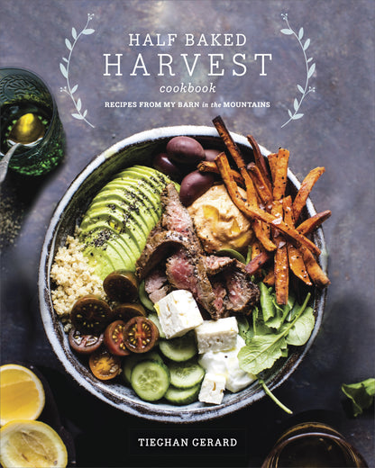 A vibrant bowl featuring sliced steak, avocado, sweet potato fries, cucumbers, tomatoes, feta cheese, quinoa, olives, and greens sits on a dark surface. This delicious comfort food recipe is showcased in the Half Baked Harvest Cookbook by Tieghan Gerard from Penguin Random House.