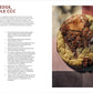 A large chocolate chip cookie with a crackled surface and melted chocolate sits on a parchment-lined baking sheet next to a printed recipe from the Cookie Club cookbook by Penguin Random House.