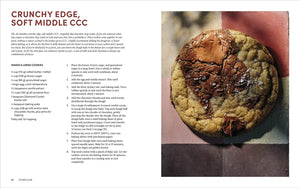 A large chocolate chip cookie with a crackled surface and melted chocolate sits on a parchment-lined baking sheet next to a printed recipe from the Cookie Club cookbook by Penguin Random House.